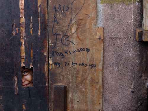 Free A playful child peeks through a wooden board, surrounded by rustic textures in an outdoor setting. Stock Photo