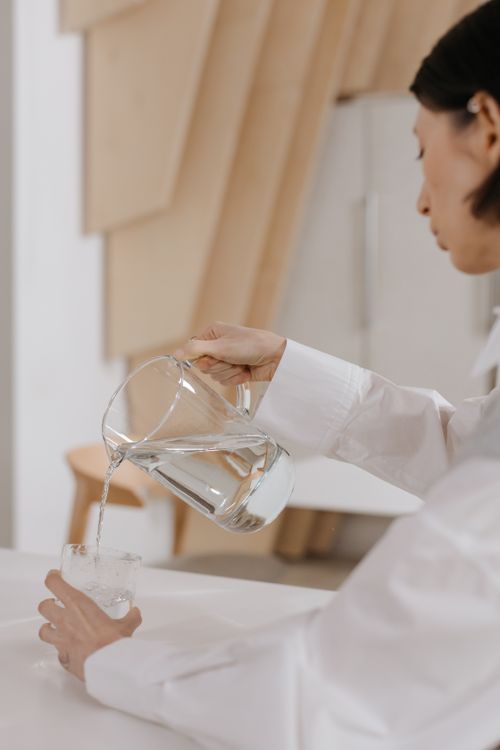 Free Woman in white shirt pouring water from a pitcher into a glass in a modern indoor setting. Stock Photo