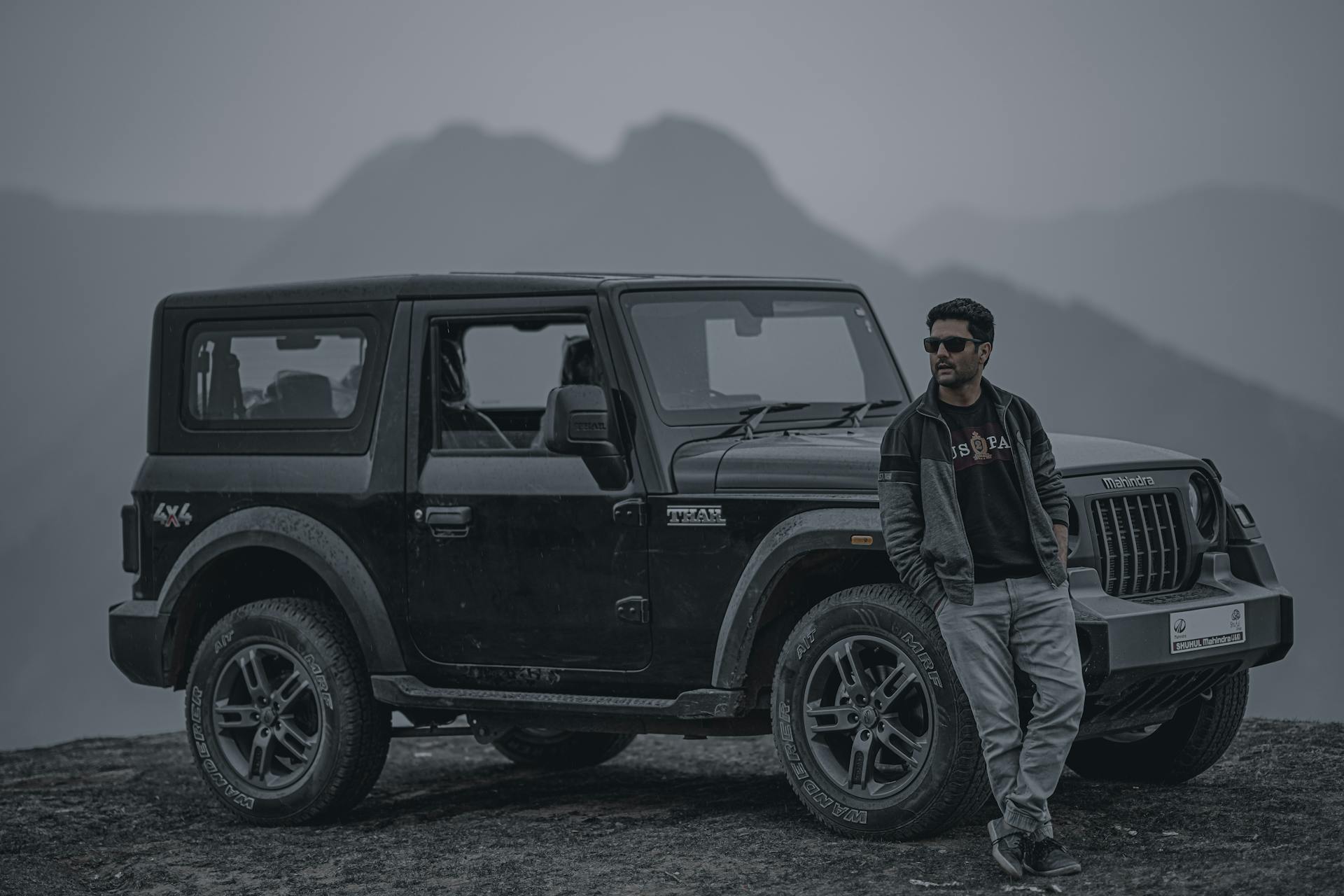 A man in a black jacket and sunglasses leans on a Mahindra 4x4 jeep, set against a misty mountain landscape.