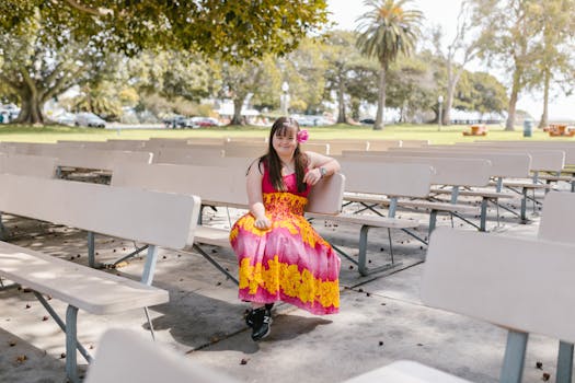 A smiling woman in a vibrant dress sits alone on a park bench surrounded by trees and greenery.