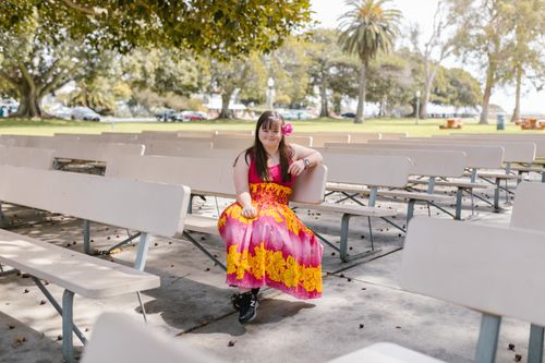 Free A smiling woman in a vibrant dress sits alone on a park bench surrounded by trees and greenery. Stock Photo