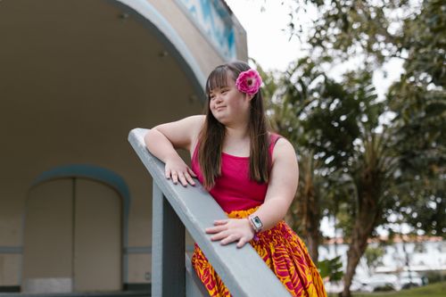 Free A young girl with Down syndrome smiles while leaning against a handrail outdoors, wearing a vibrant floral outfit. Stock Photo