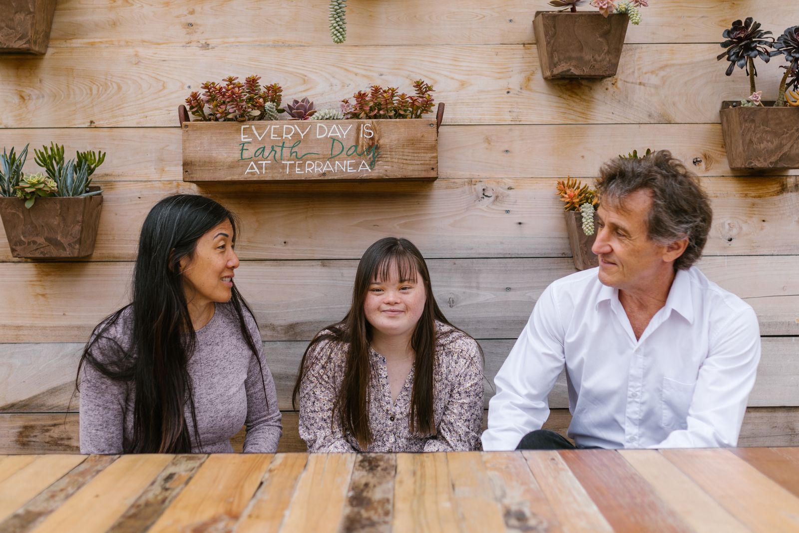 An adult with Down syndrome sits and talks together with two smiling family members outdoors.