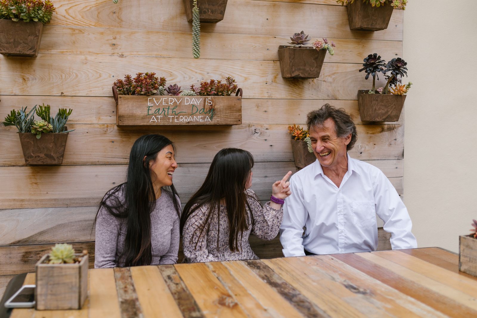 An adult with Down syndrome laughs together with two family members at an outdoor table.