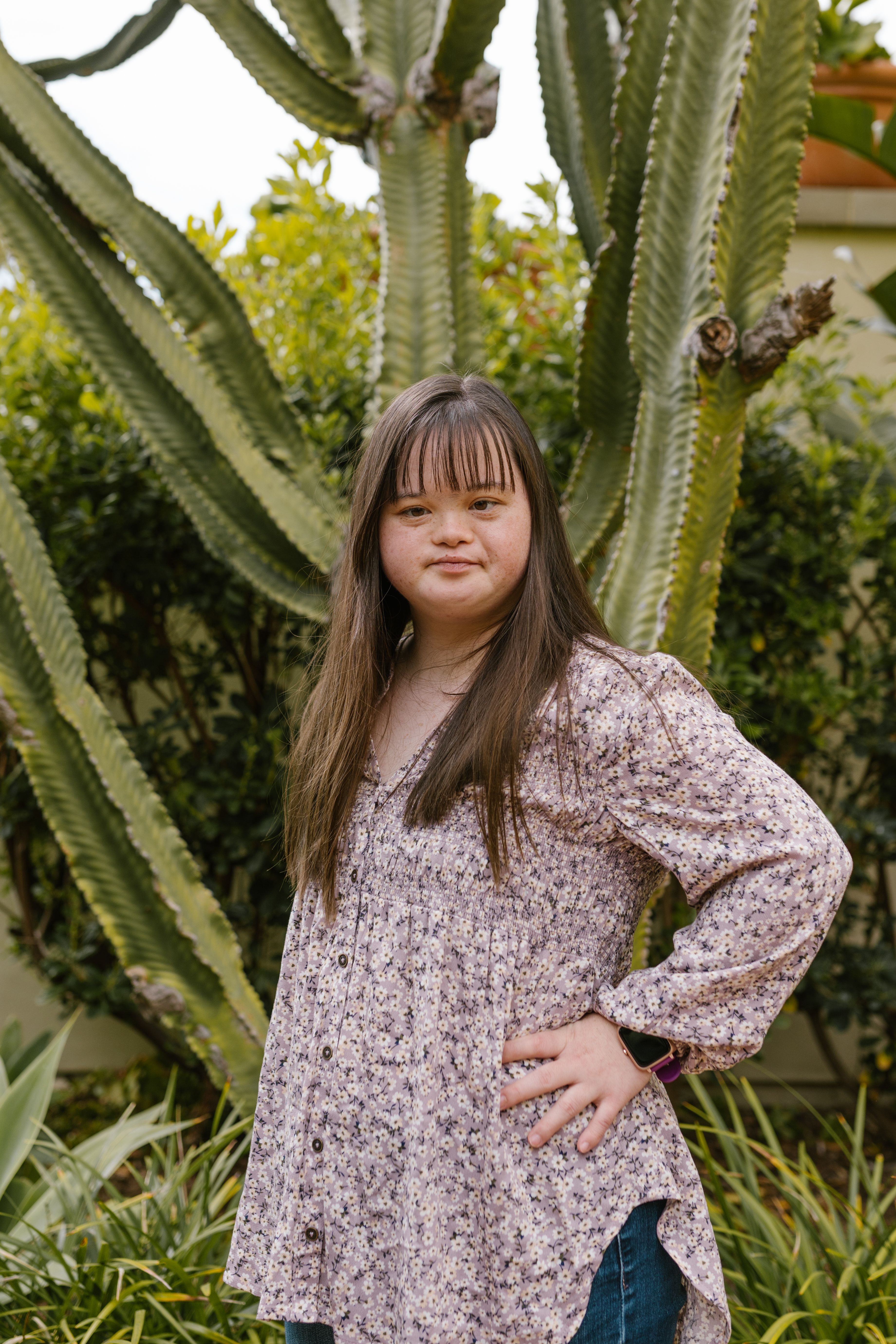 Young woman wearing floral top posing confidently in a lush garden, featuring large cacti.