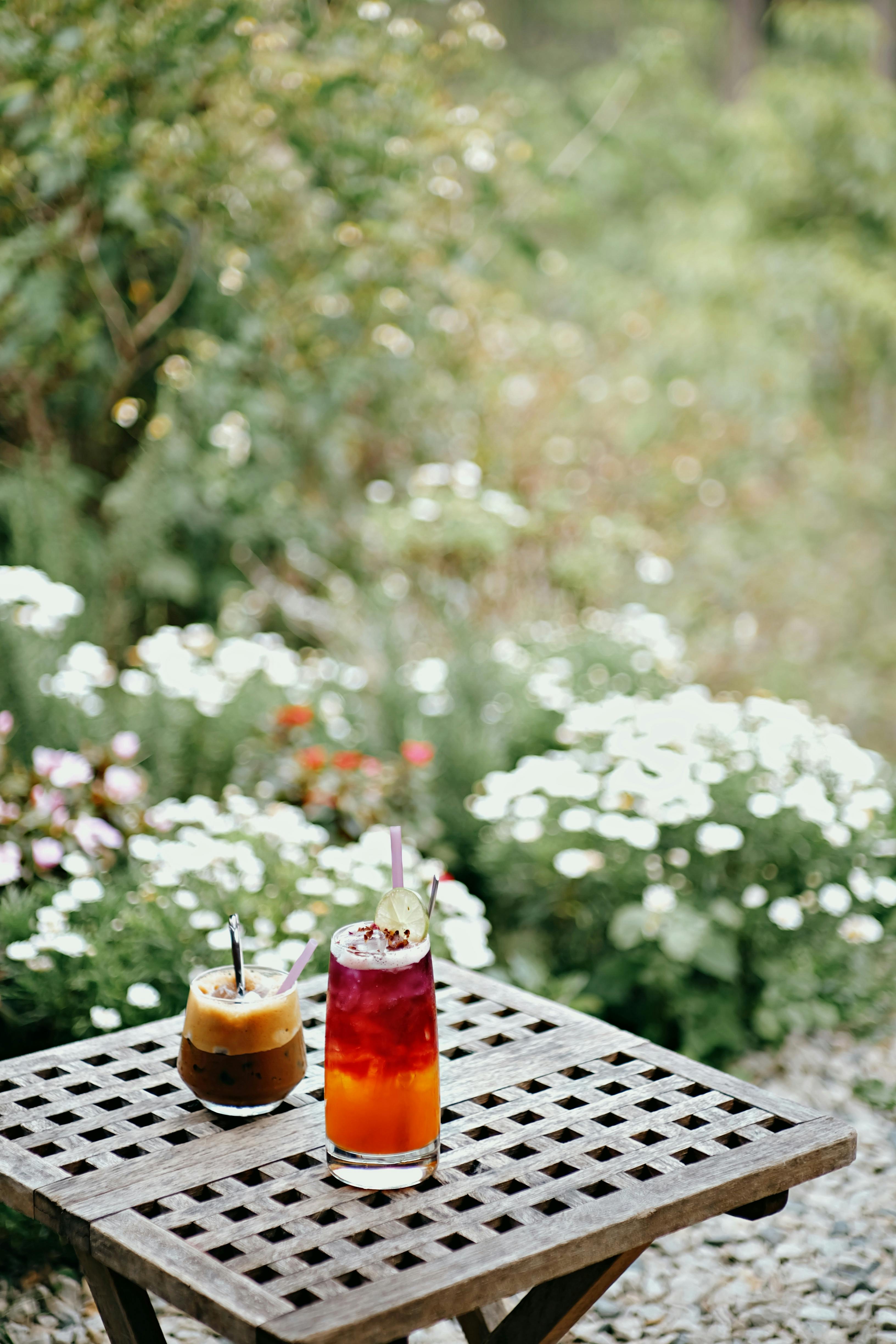 Two iced drinks on a checkered table amidst a lush garden backdrop with flowers.