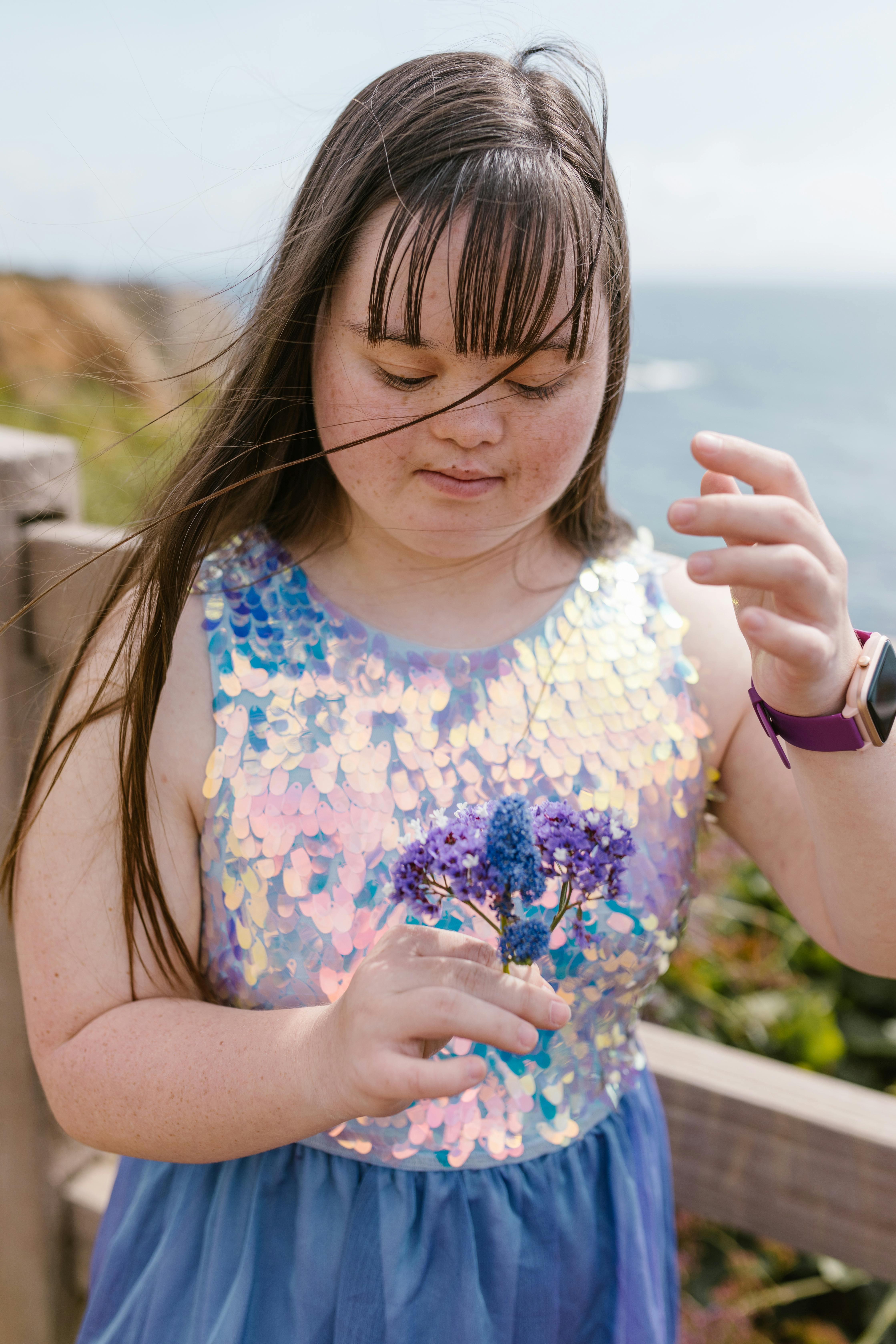 Young girl with Down syndrome in a sparkling dress holds purple flowers outdoors by a wooden fence.