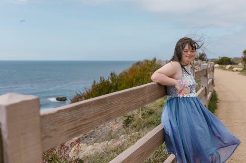 A Girl in a Blue Dress Leaning on a Wooden Fence