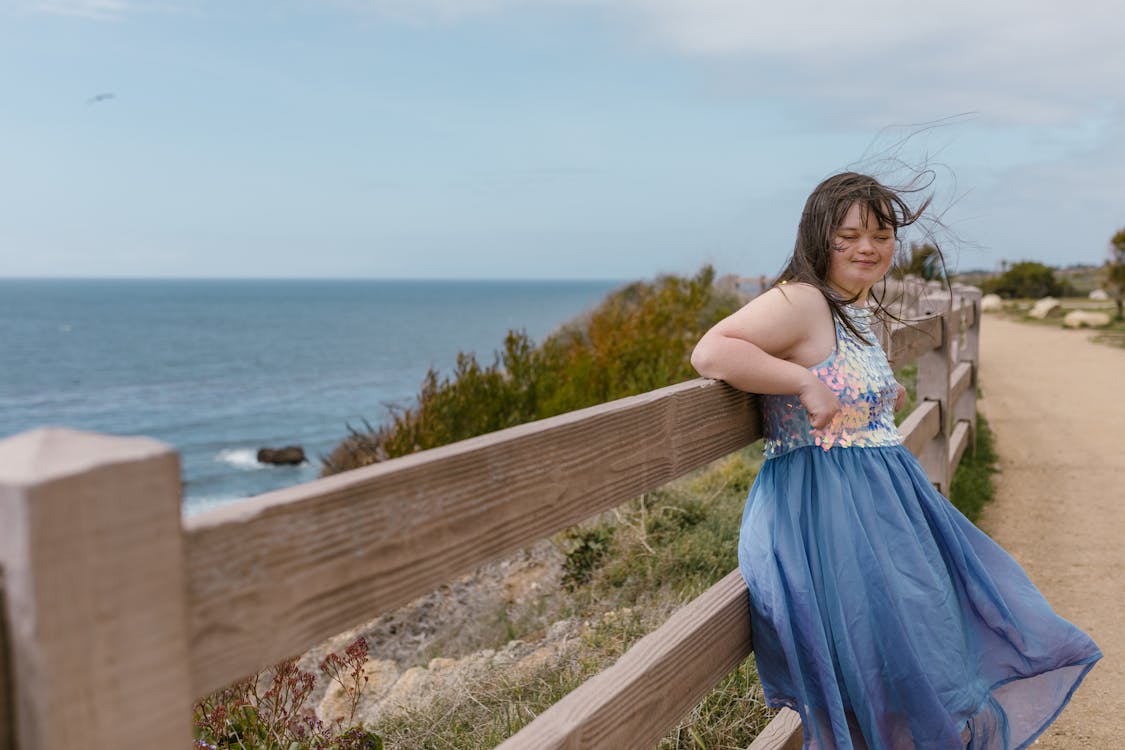 A Girl in a Blue Dress Leaning on a Wooden Fence