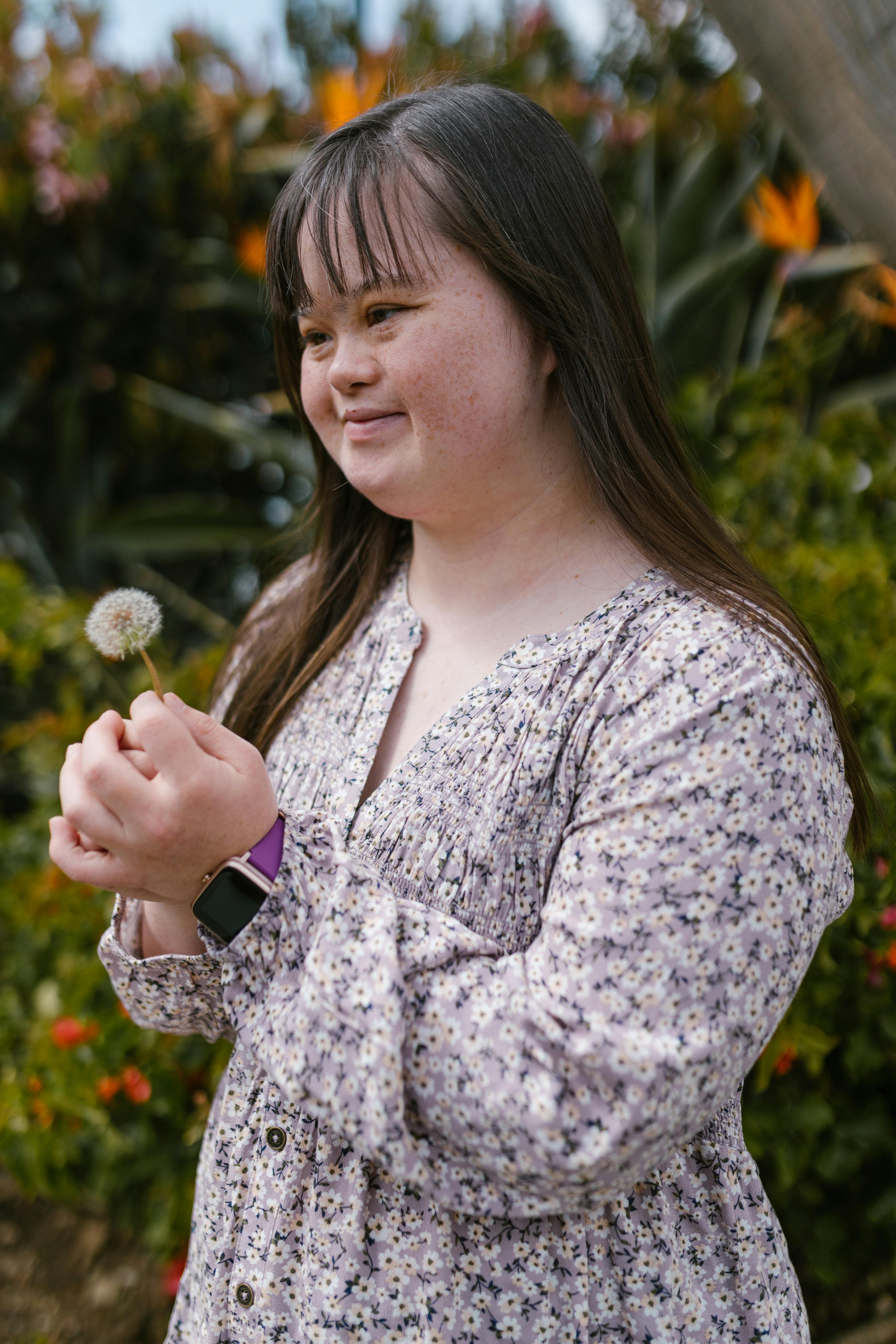 A young woman with Down syndrome holds a dandelion in a vibrant garden, wearing a floral top.
