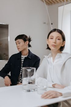 Two people in a modern, minimalist setting, exchanging thoughtful looks while sitting at a table with water pitchers.