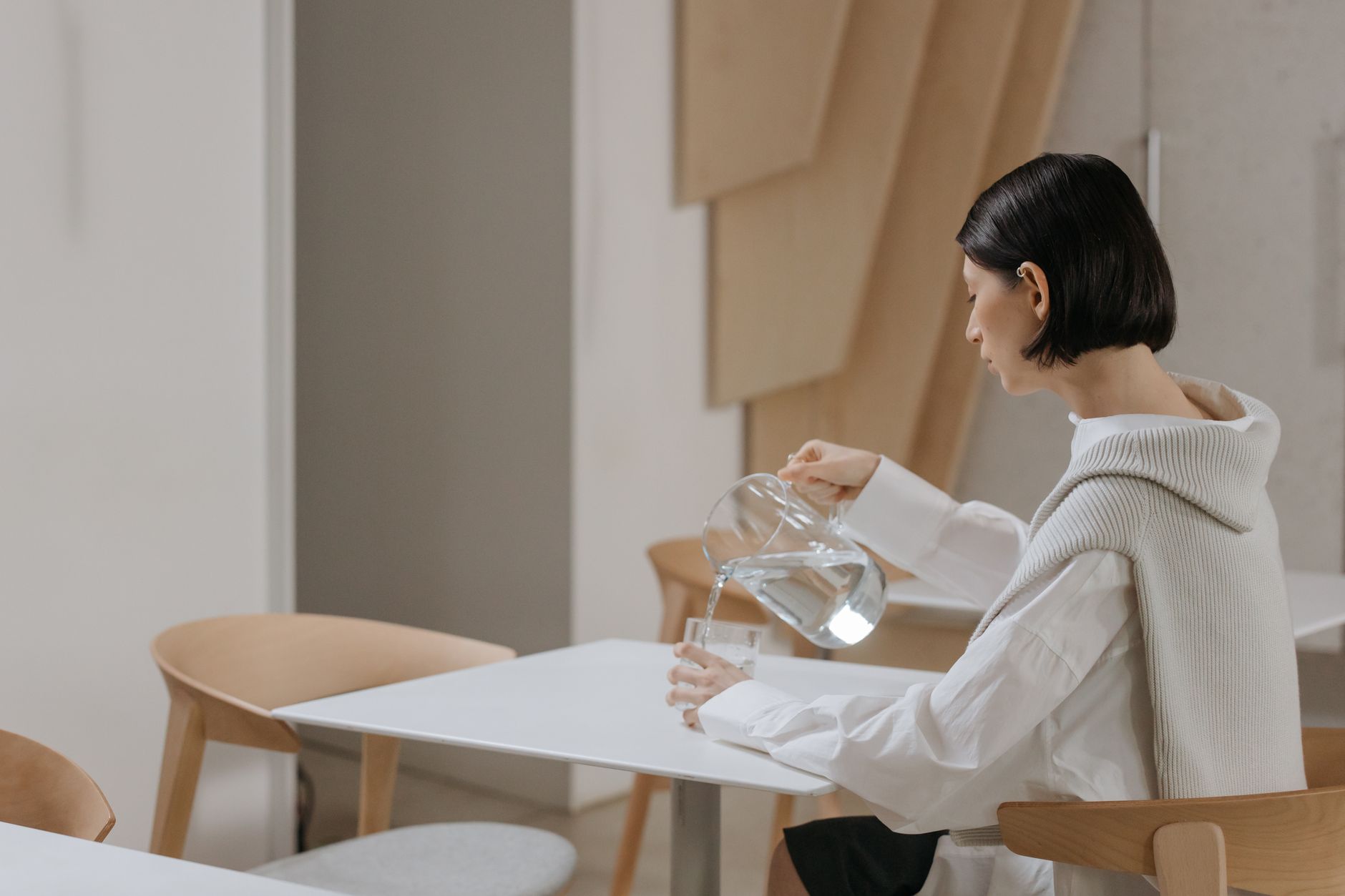 Adult woman in white pouring water into a glass on a minimalist table indoors.
