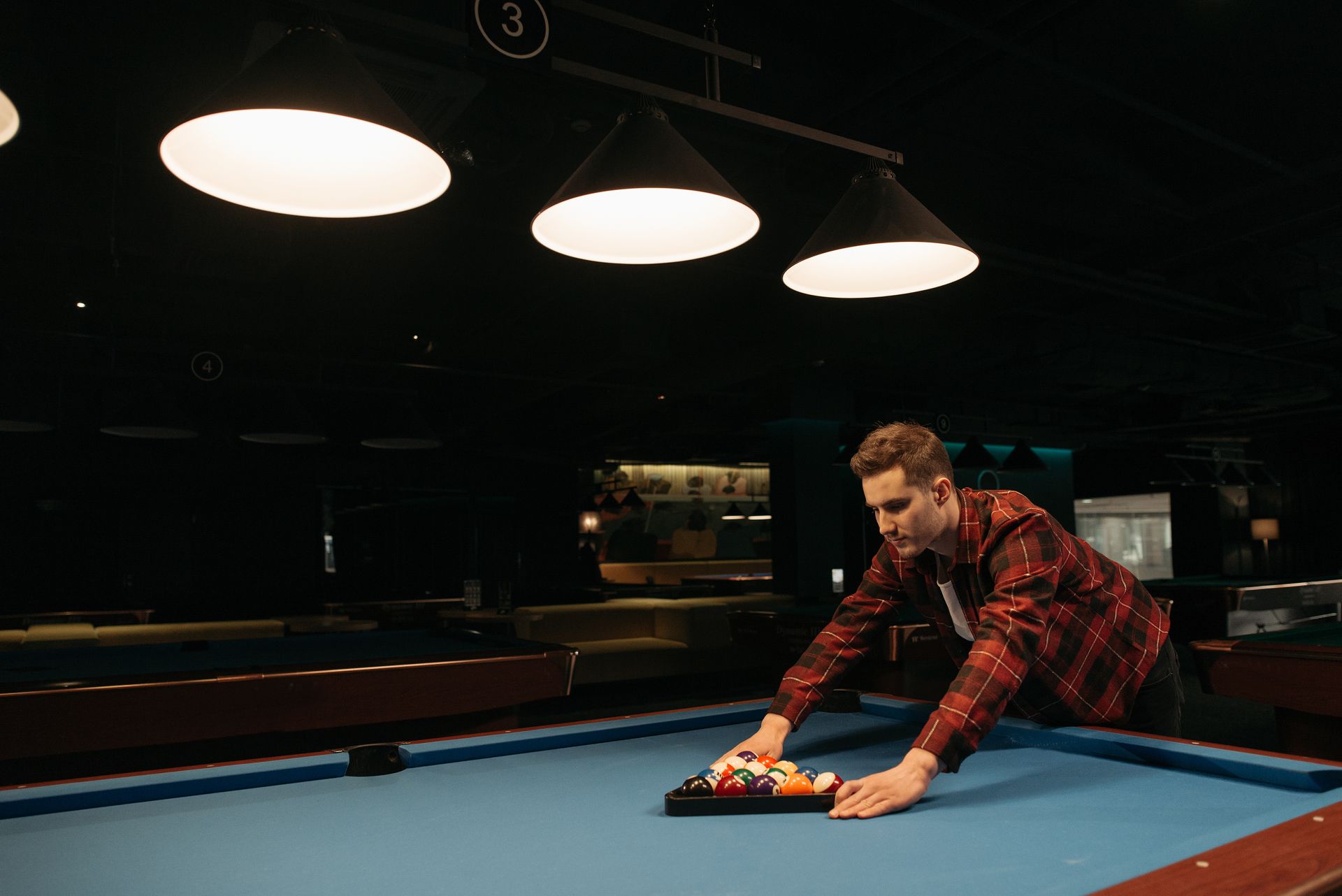 A man setting up billiard balls on a pool table in a dimly lit billiard hall.