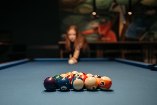 Free A close-up of billiard balls on a blue table with a woman preparing to play in the background. Stock Photo