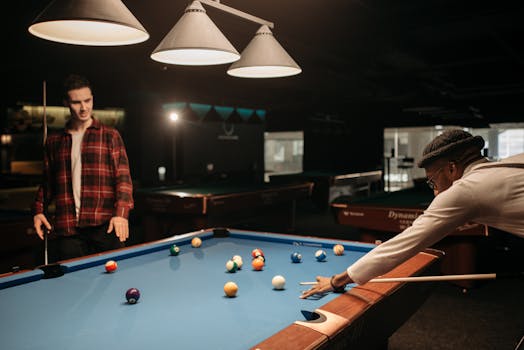 Two men playing pool on a blue table in a dimly lit room, enjoying a leisure activity.