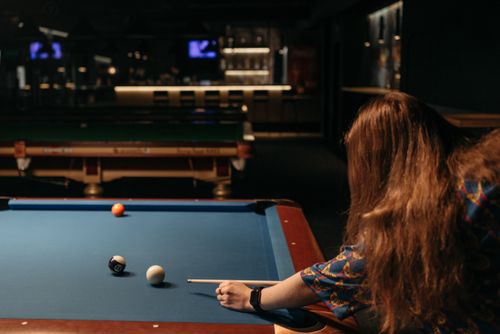 Free A woman with red hair playing billiards in a dimly lit pool hall, aiming with a cue stick. Stock Photo