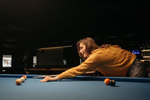 Free Woman in a yellow sweater playing billiards on a blue table in a dimly lit indoor setting. Stock Photo