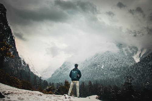 Full body back view of anonymous backpacker with rucksack contemplating mountain ridge while standing on edge of snowy mount in foggy weather