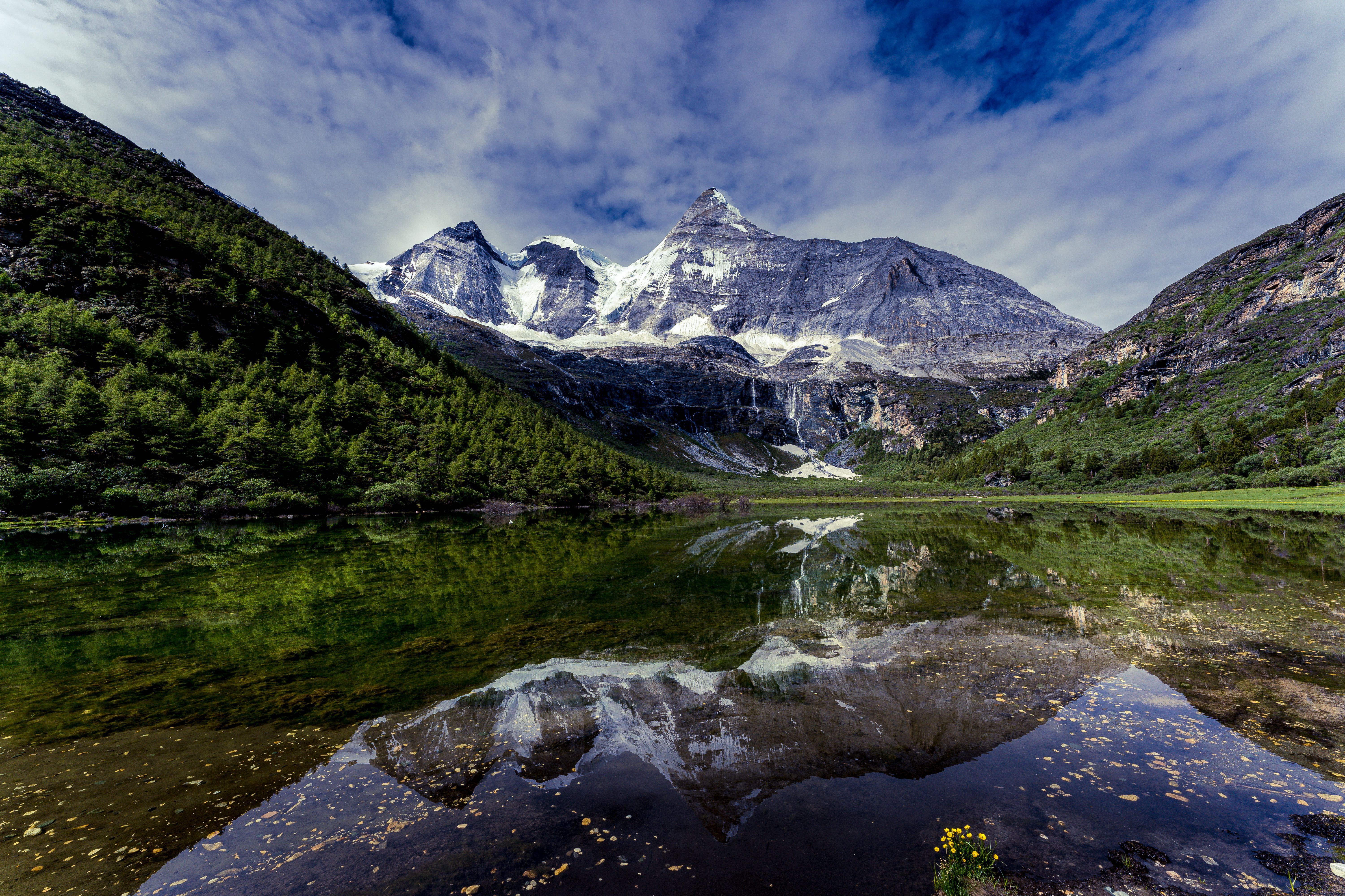 Crystal-clear lake reflecting jagged snow-capped peaks of Nianbaoyuze, with wildflowers in the foreground