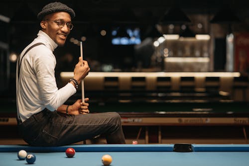 A Smiling Man in White Turtle Neck Sleeves Holding a Cue Stick while Sitting on a Pool Table