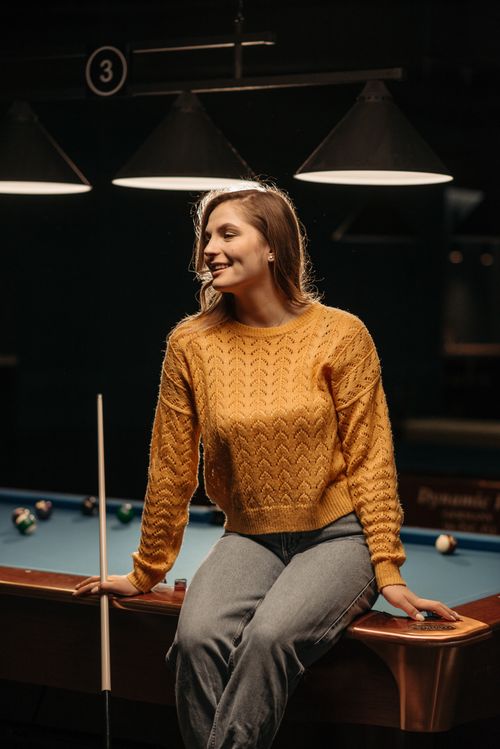 Free A woman in a yellow sweater casually sits on a billiard table with a cue stick. Stock Photo