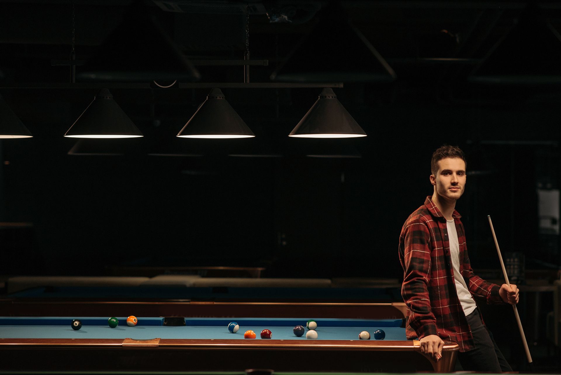 A man leaning on a billiard table holding a cue stick in a dimly lit room.