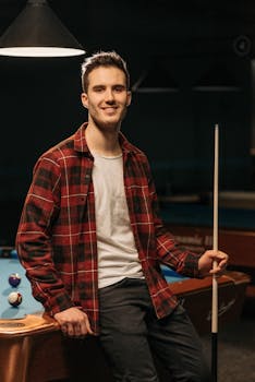 Smiling young man in flannel shirt poses with cue stick by billiard table.