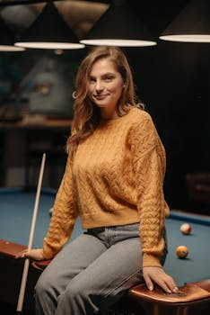 Woman sitting on a billiard table with a cue stick, wearing a brown sweater in a warm, cozy setting.