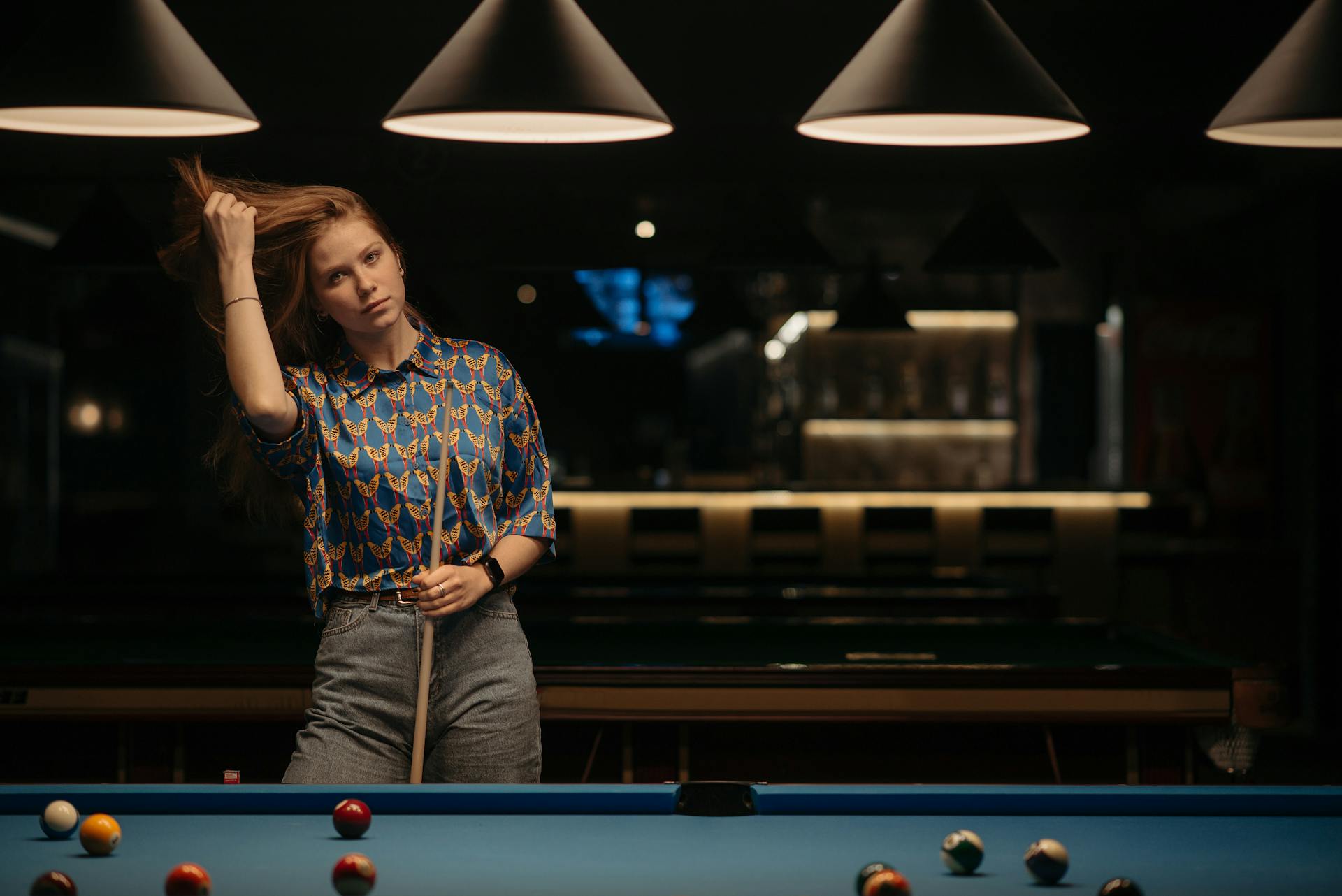 A woman stands by a pool table in a billiard hall, holding a cue stick.