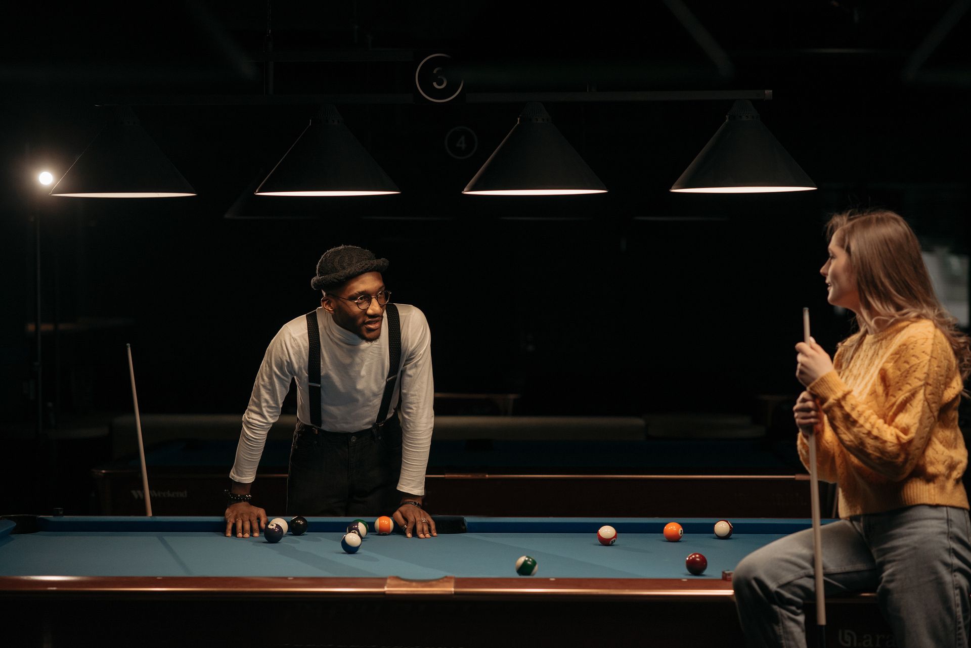 Two people enjoying a game of billiards under dim lighting in a pool hall.