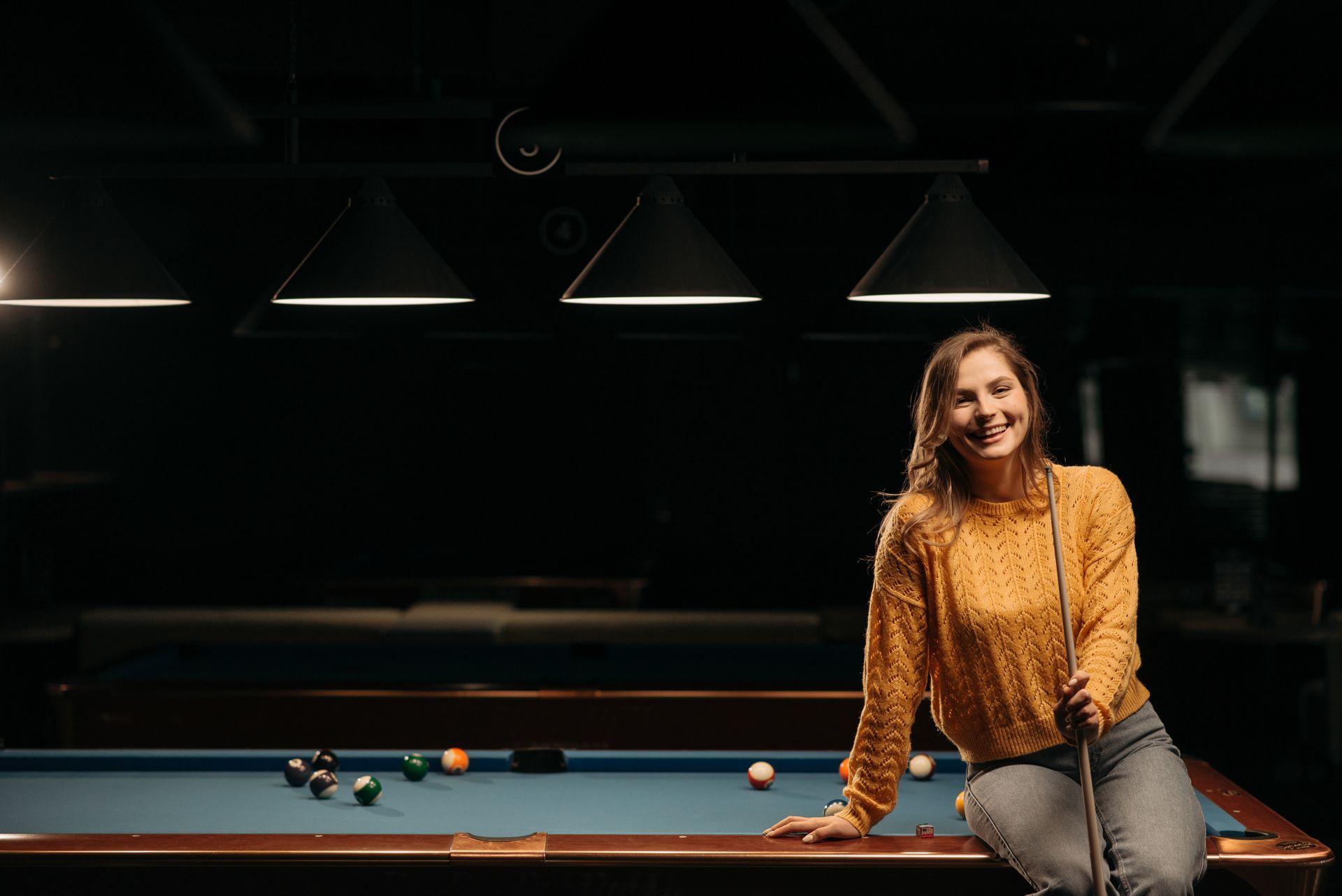 Happy woman in a yellow sweater sitting on a billiard table with cue stick in a cozy indoor setting.