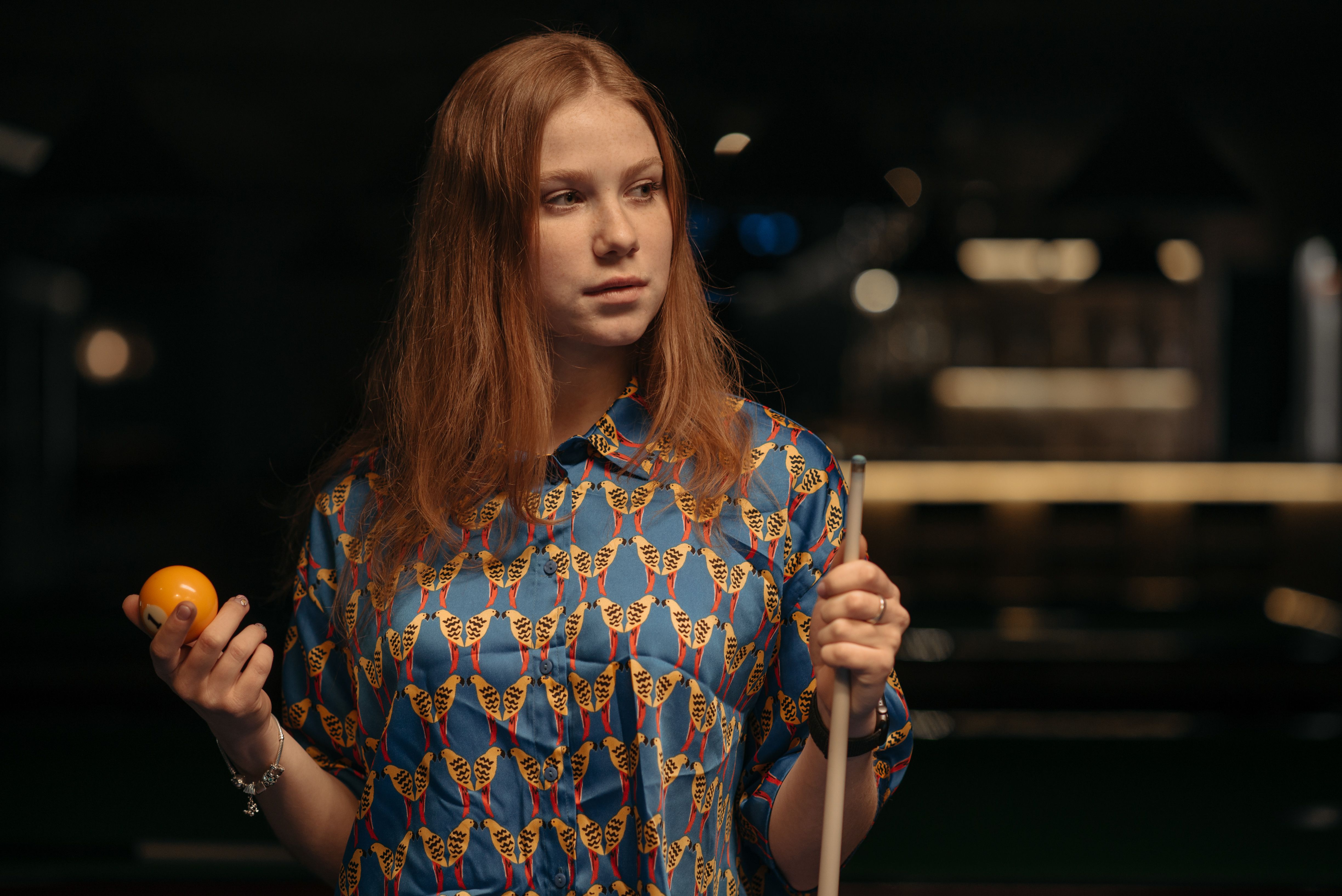 A woman in a colorful shirt holds a billiard ball and cue stick, poised for a game indoors.