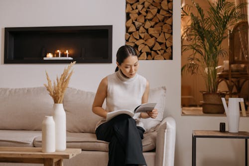 A Woman in White Turtleneck Top Sitting on the Couch while Reading a Book