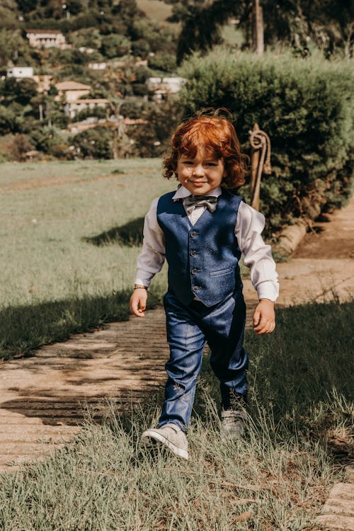 Smiling child in formal wear with red hair looking at camera while strolling on pathway in sunlight
