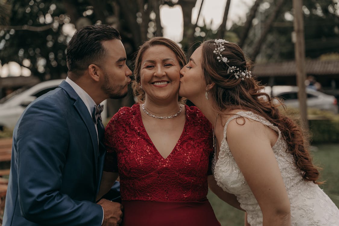Ethnic bride and groom kissing smiling mom in elegant wear during festive event in countryside