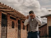 Stylish ethnic man putting on sunglasses against old building