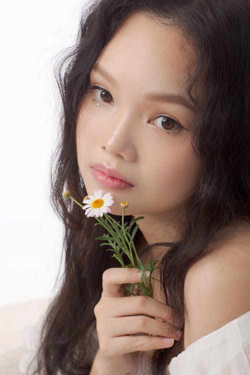 Asian female with makeup touching face gently with chamomile and looking at camera thoughtfully on white background