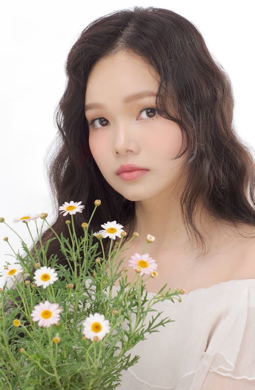 Wavy haired Asian woman holding chamomile flowers and looking at camera against white background