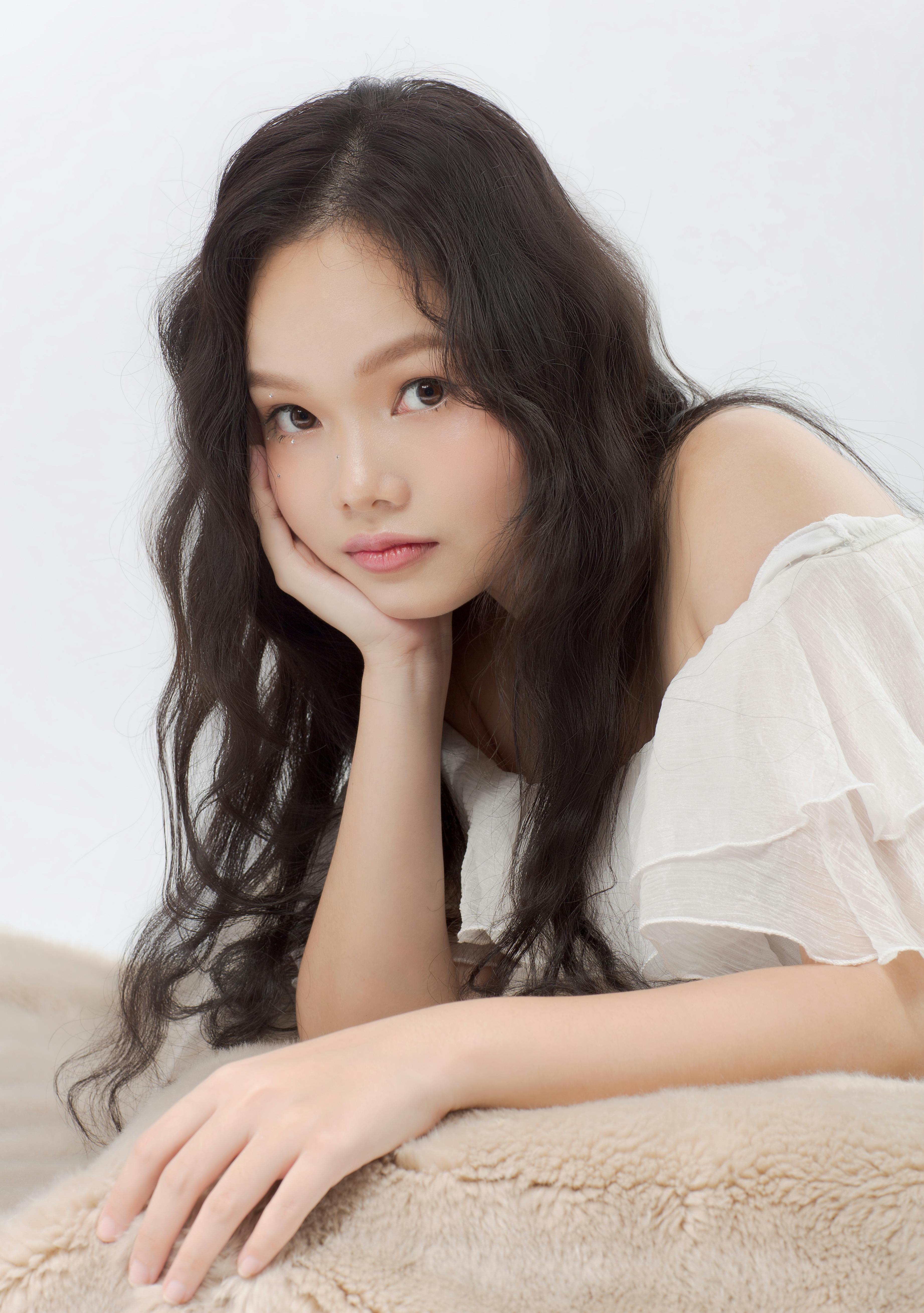 Long haired Asian female in white outfit looking at camera while leaning on hand on fur plaid on white background