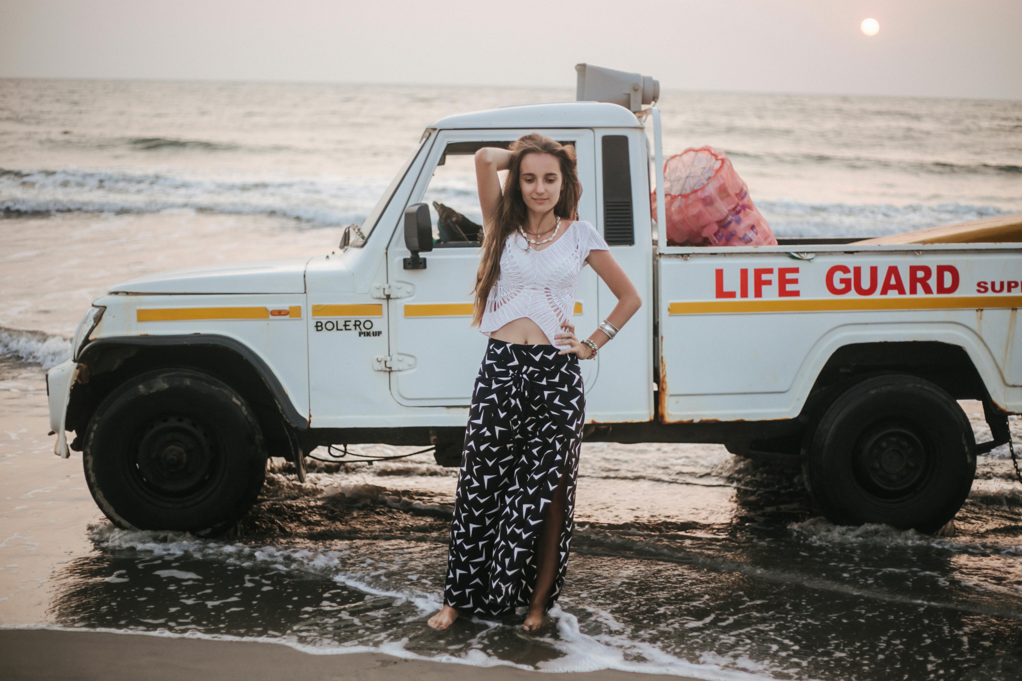 A fashionable woman poses by a lifeguard truck at sunset on a beach, showcasing summer vibes.