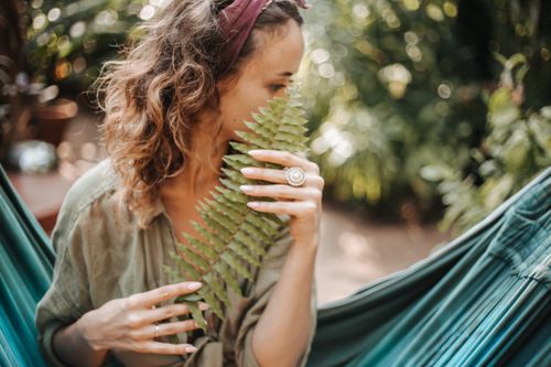 Free A woman enjoys a tranquil moment in a hammock, holding vibrant green fern leaves outdoors. Stock Photo