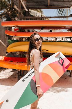 Smiling woman in sunglasses holding a colorful surfboard on a sunny beach day.