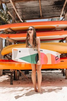 Smiling young woman holding a surfboard on a sunny beach with colorful surfboards.