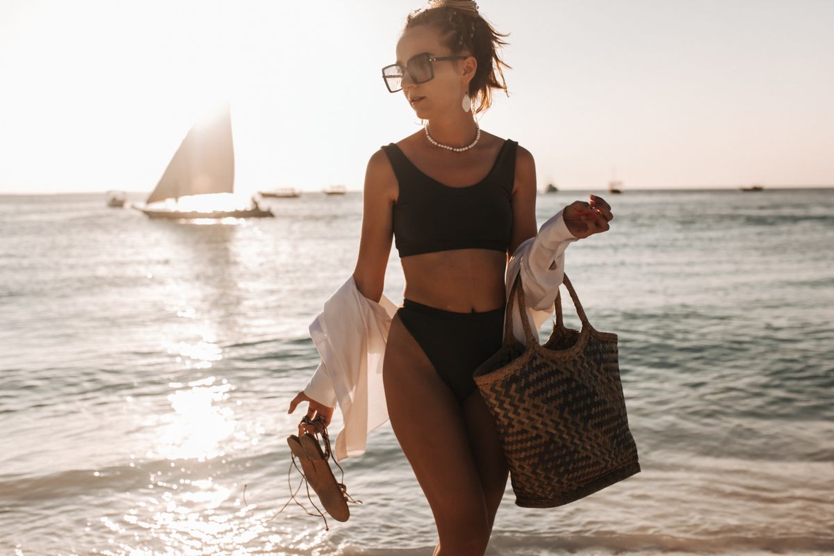 A woman in swimwear carrying a bag on a beach — styling the swimsuit with accessories