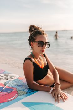 Stylish woman in swimwear and sunglasses relaxing on the beach with a surfboard.