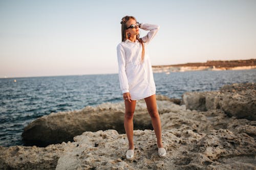 Free A Woman in a White Dress Standing on a Rocky Coast Stock Photo