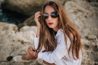 自信的女人戴着墨镜和白色连衣裙坐在海滩的岩石上