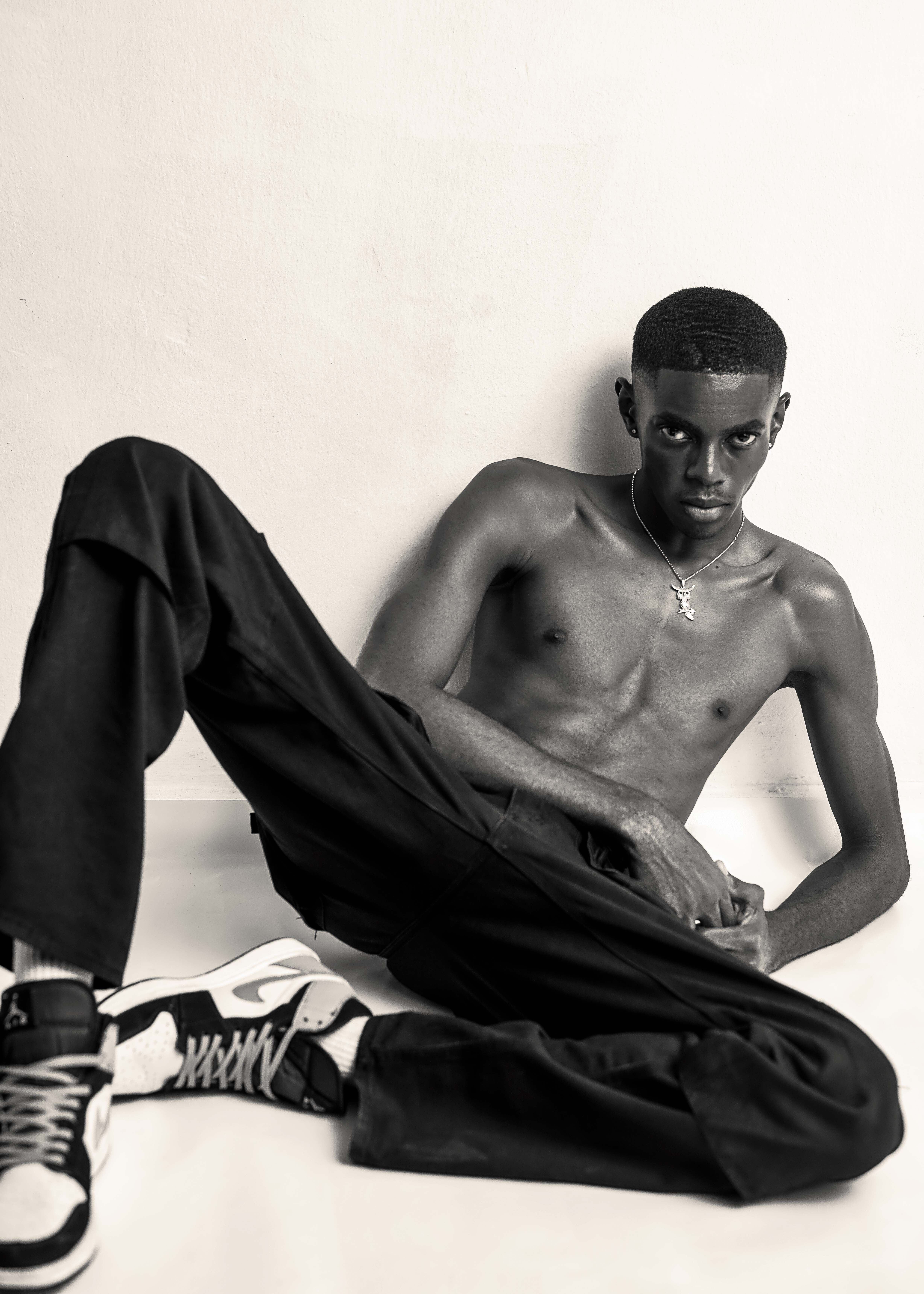 Minimalist portrait of a shirtless man sitting on a white backdrop, wearing black pants and sneakers.