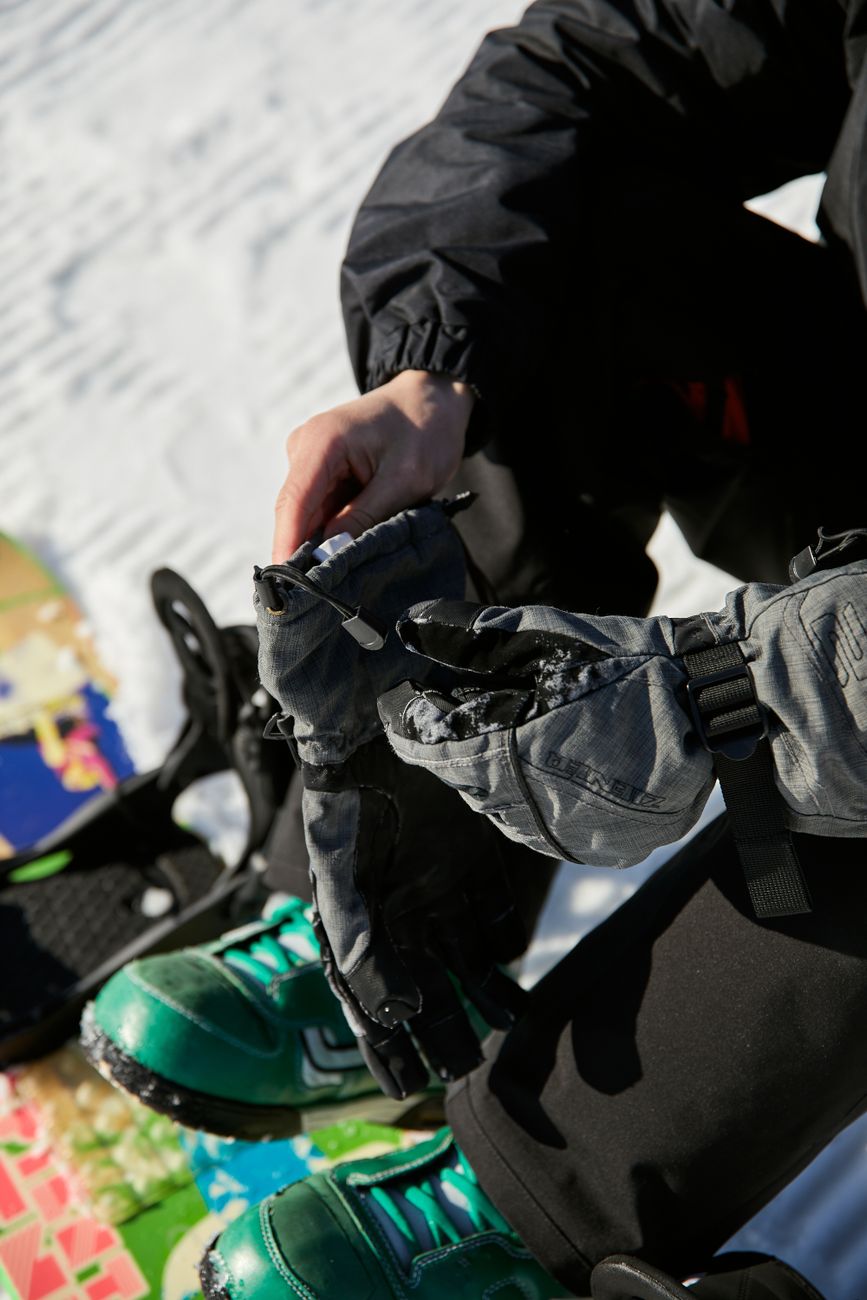 Person sitting on snow with snowboard, adjusting gloves in bright winter daylight.