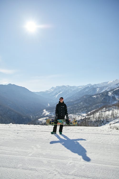A Man in Black Jacket and Pants Standing on a Snow Covered Ground while Holding a Snowboard
